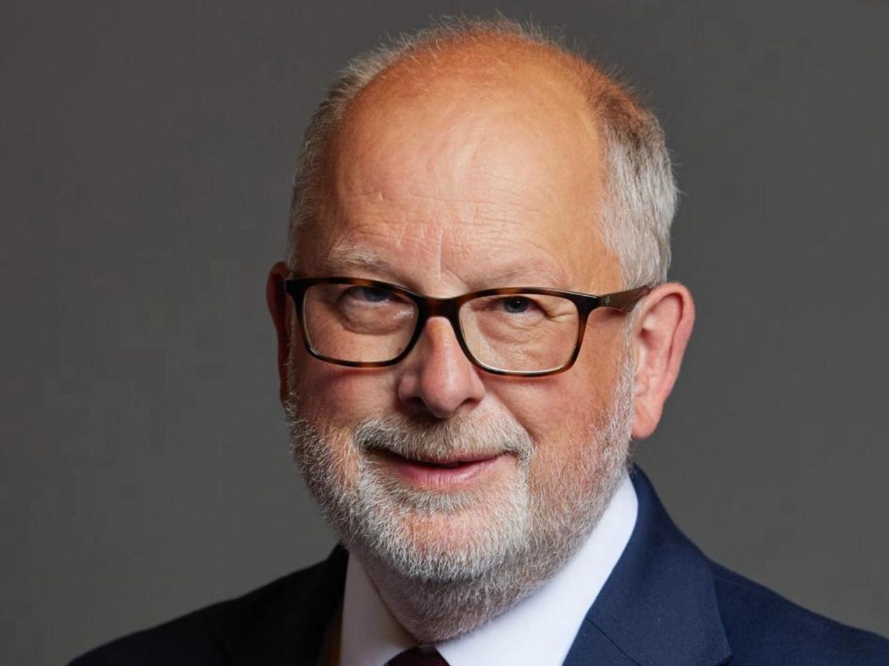 A man wearing a dark blue suit. He's wearing glasses and has facial hair and a beard