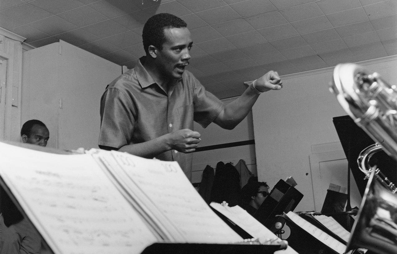 Quincy Jones gestures as he interacts with a brass section in a recording studio, that are just out of view save for a little bit of saxophone, with some music notation on a music stand in front of the saxophone. Picture is taken in 1963.