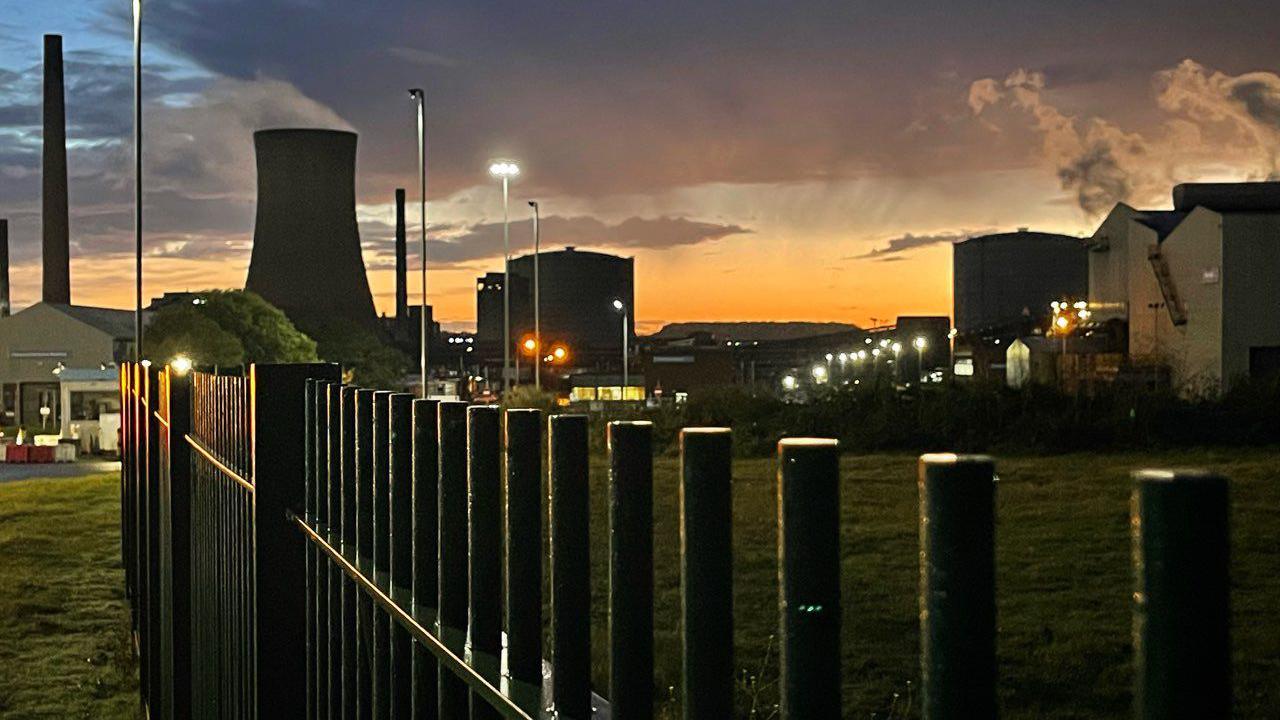 Steel works at dawn, the sky is clouded and orange with the buildings in silhouette.