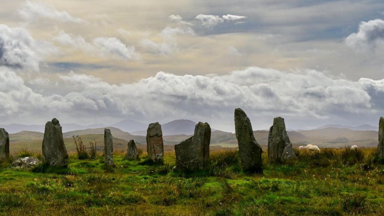 Landscape image of the Calanais Standing Stones - around 10 stones are visible, about 5 meters tall. There are clouds overhead and the grass is green with a yellow tint, showing the change of the seasons