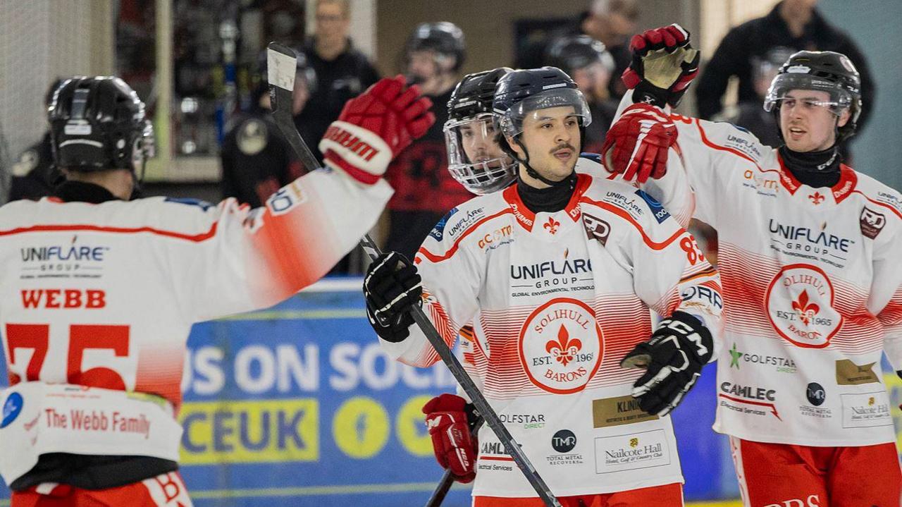 Three men in white ice hockey jerseys with red lettering, wearing black helmets, celebrating together on an ice rink