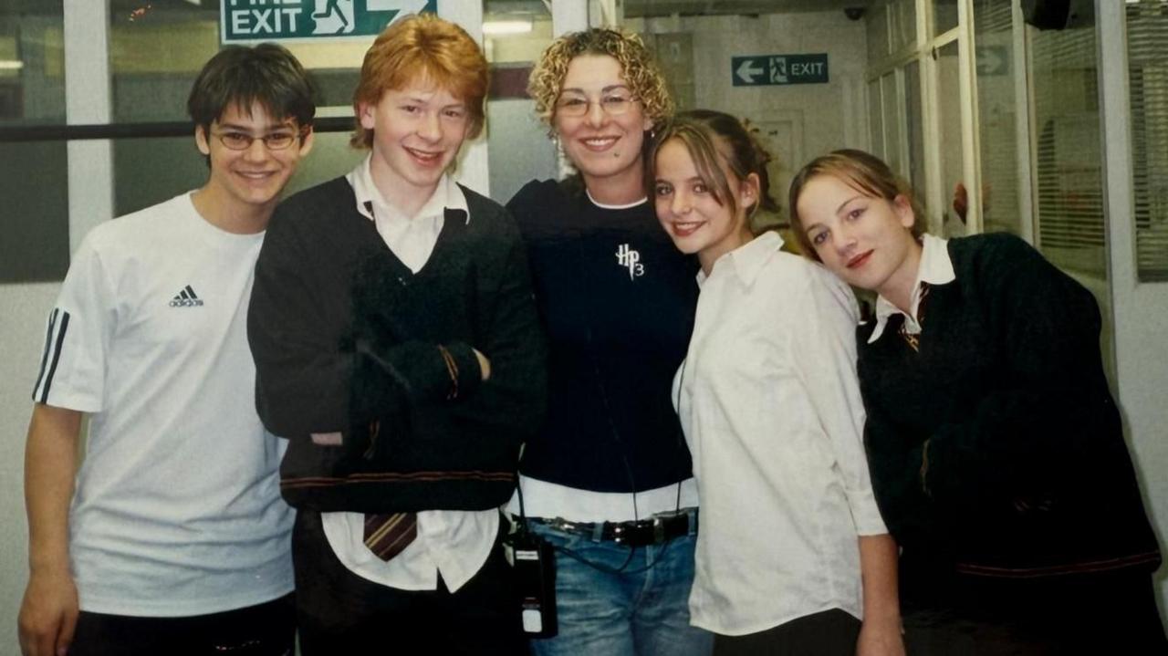 Five people standing in a hallway. They are dressed in casual clothes and school uniforms, including shirts, jumpers and ties.