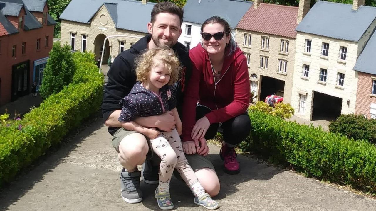 Luna, Louis and Natasha. They are at what appears to be a model town, with littl model buildings around them in the background. Both adults are squatting down, holding hands. Louis has his arm around Luna, who has leggings with dogs on, a dotted hoodie, and is half sitting on his knee. Natasha has sunglasses on and her hair in a ponytail. All three are smiling at the camera. 