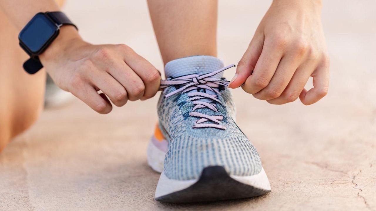 A close-up of a blue and white trainer with pink laces being tied up. The person tying them up is wearing a black watch.