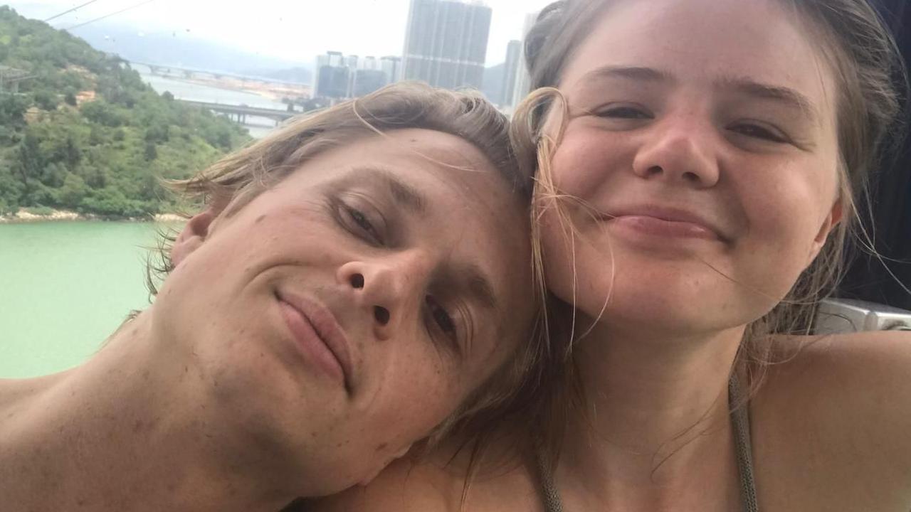 A couple look at the camera for a selfie. They appear happy and smiling. The man has blond hair and is leaning on the shoulder of the woman who has fair hair which is tied back from her face.