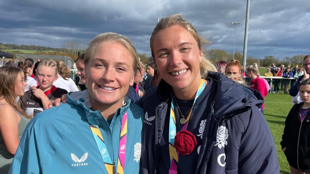 Red Roses Alex Matthews and Zoe Aldcroft in front of a pitch filled with young female rugby players.