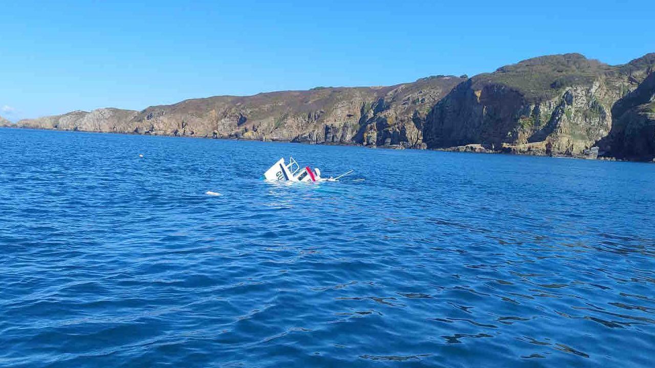 Part of a white boat called Wild Wave pokes out of the surface of the sea water. The bow of the boat is white, blue and red. The water is calm. A cliff edge is in the background.