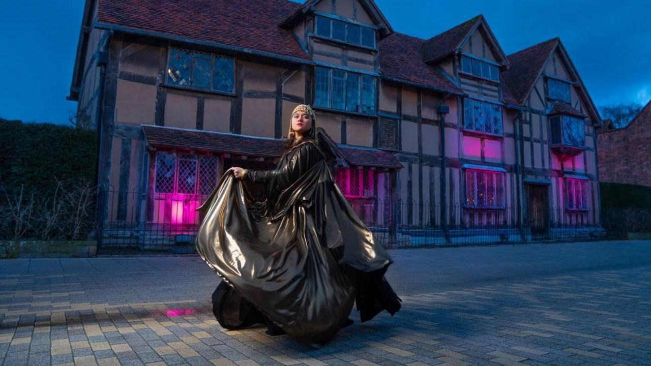 A woman in large black dress and hat with gold trim, holding up her dress on a cobbled street in front of a timber framed building which is illuminated in purple light.