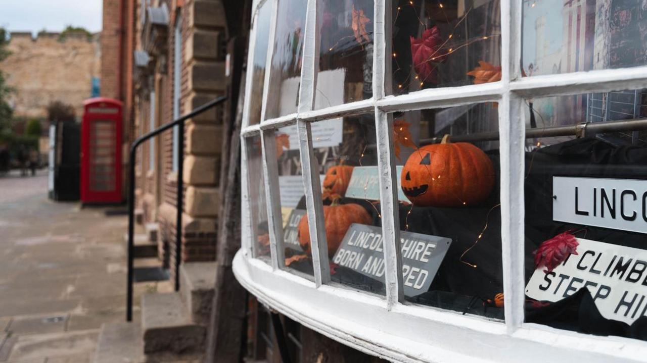 Side view of a shop window of three orange pumpkins with faces and signs such as 'Lincolnshire born and bred'. A telephone box can be seen in the distance and Lincoln Castle wall.