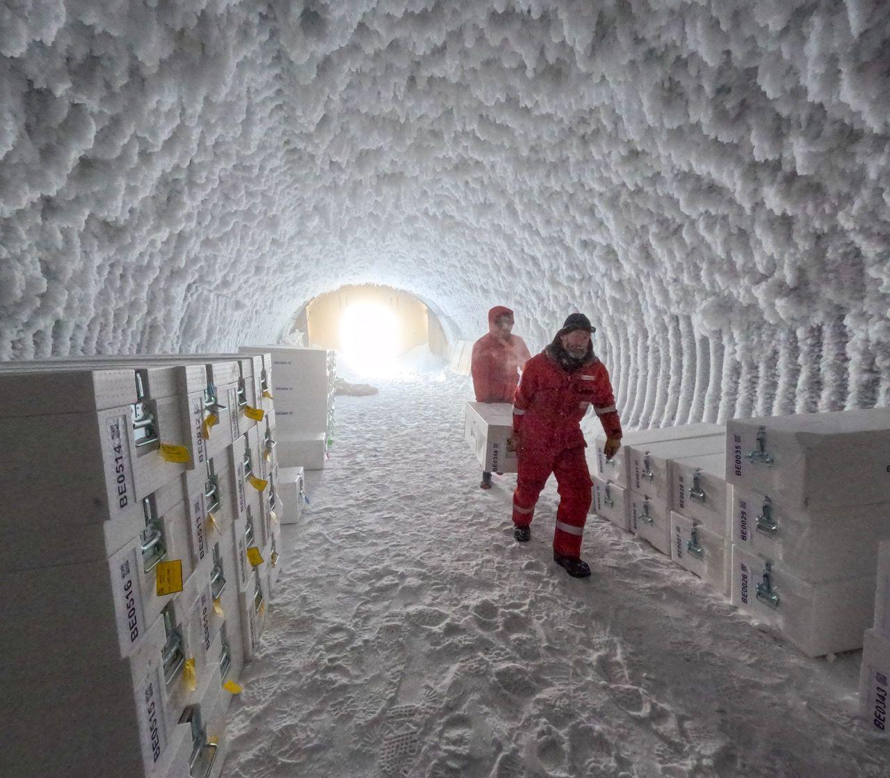 two people in sub zero protective red jump suits carry a large white chest between them through an icy tunnel towards the camera. Lots more similar boxes are stacked on each side of the walls of the cave.