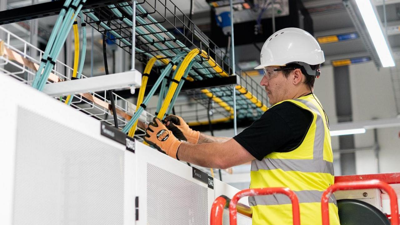 An engineer standing on a raised platform works on wiring in a datacentre.