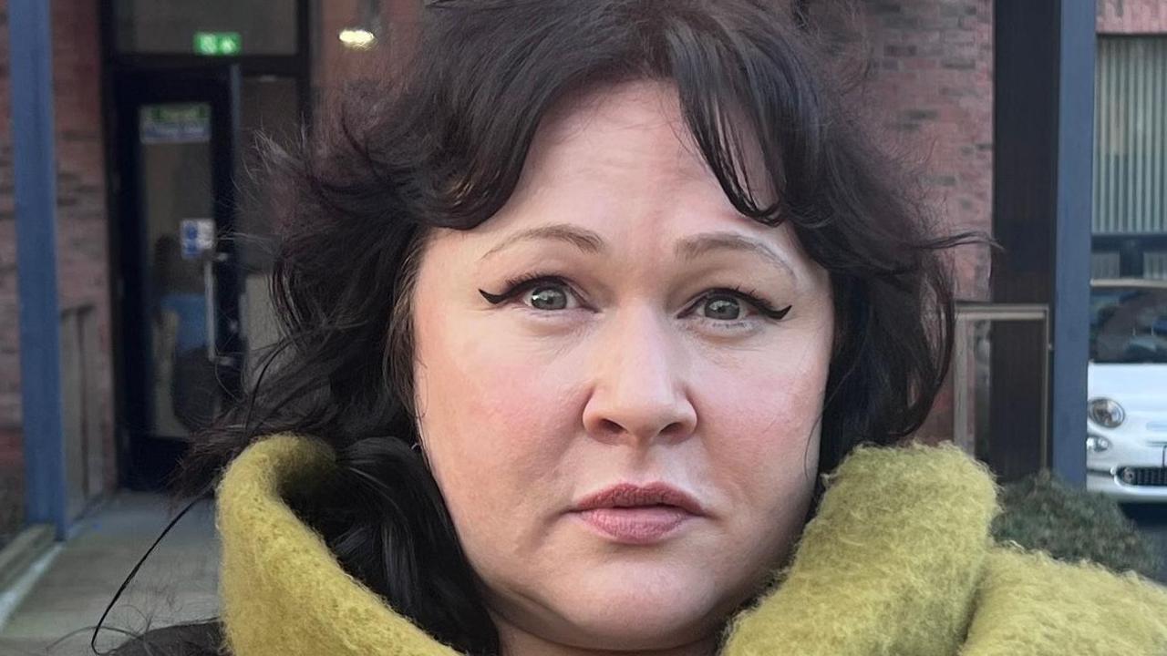 Ms Morrison has dark black hair and is wearing makeup including a winged eyeliner. She has a bright green scarf and a black top.