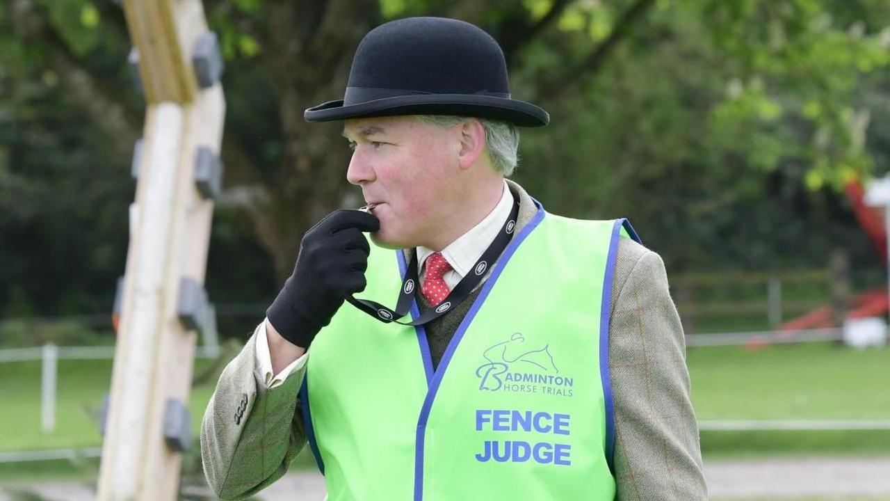 Martin wears a high-vis fence judge jacket and a bowler hat. He is blowing a whistle.