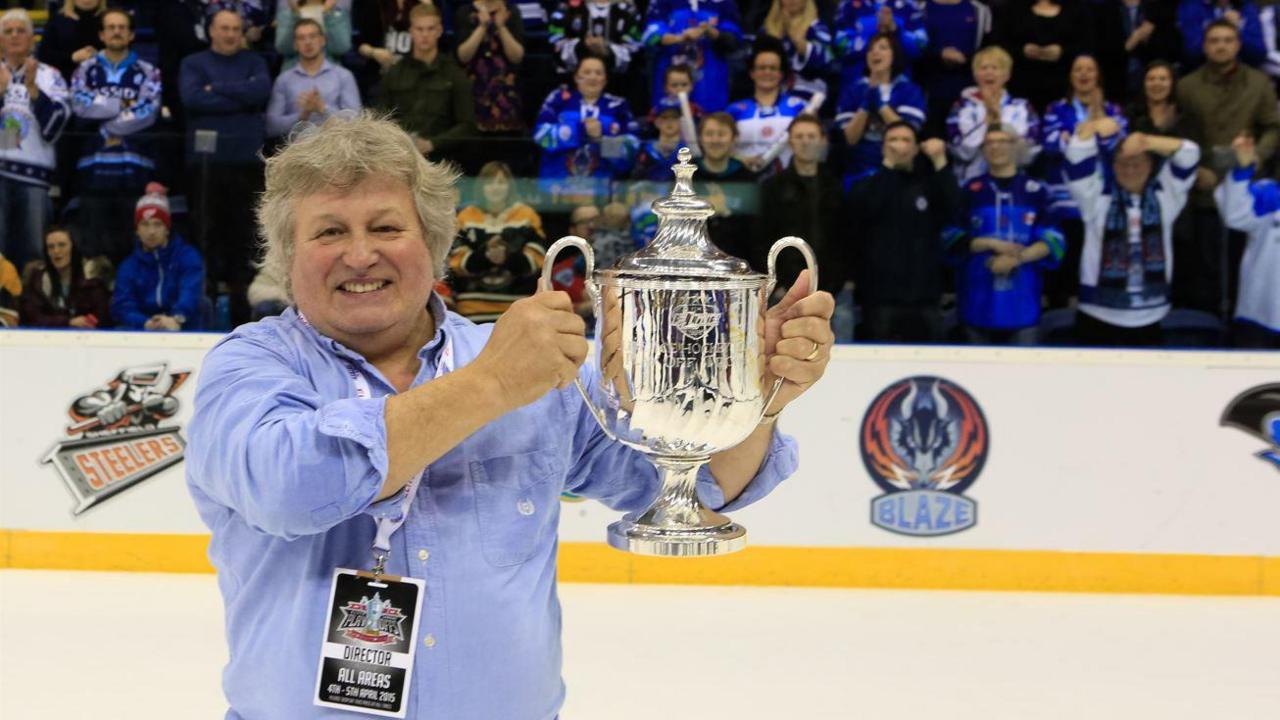 A smiling man with grey hair and a blue shirt standing on an ice rink holding a silver trophy with both hands, with fans wearing blue clothing behind him