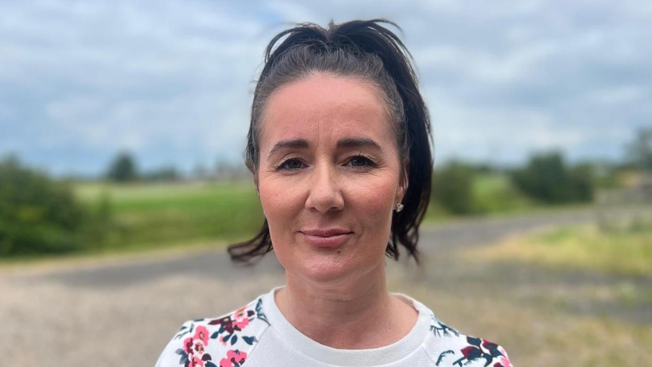 Josephine Allen standing by a road, with fields in the background. She is looking to the camera and is wearing a white jumper with floral design on the shoulders and has black hair.