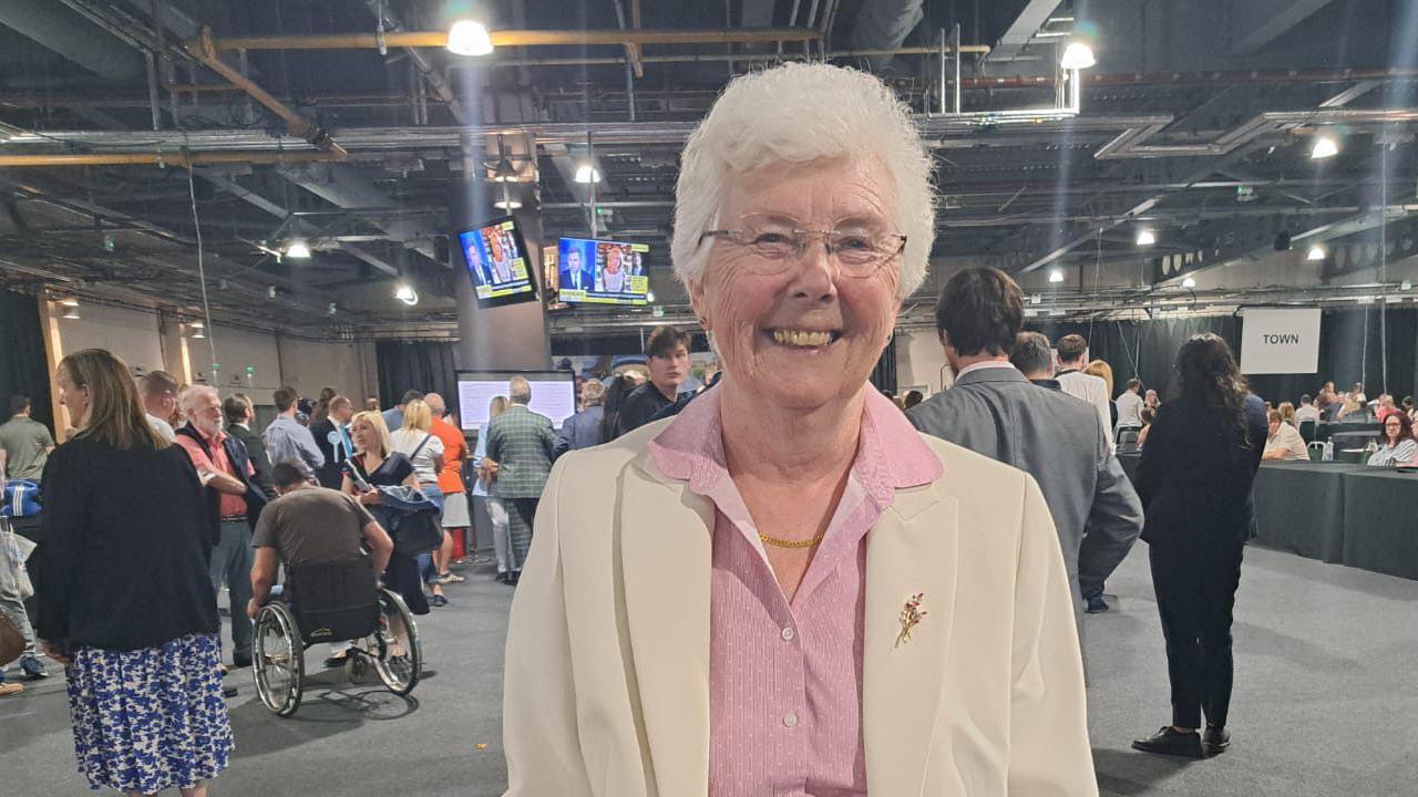 A woman with short white curly hair and glasses smiles at the camera. She is wearing a pink blouse under a cream jacket. She stands in a large conference type hall with TV screens in the background and people standing behind her looking at the screens.