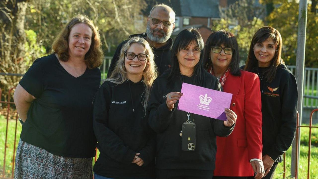 Six people are standing together in front of railings looking at the camera and trees and bushes are behind them.