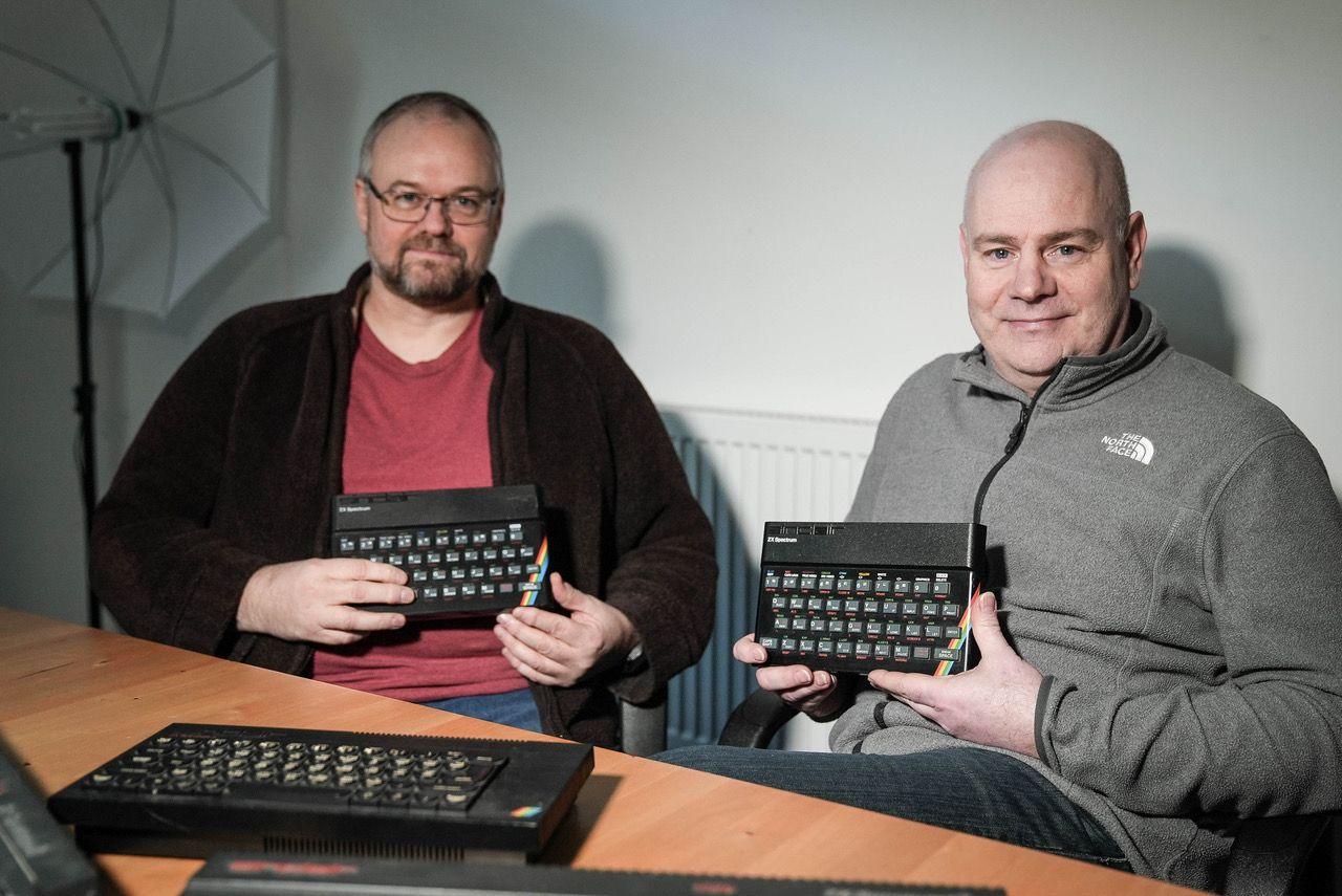 Two men in their 50s sitting at a table holding ZX Spectrum home computers. One man has glasses and a red Tshirt, the other is wearing a grey hooded top. Another ZX Spectrum is on the table 