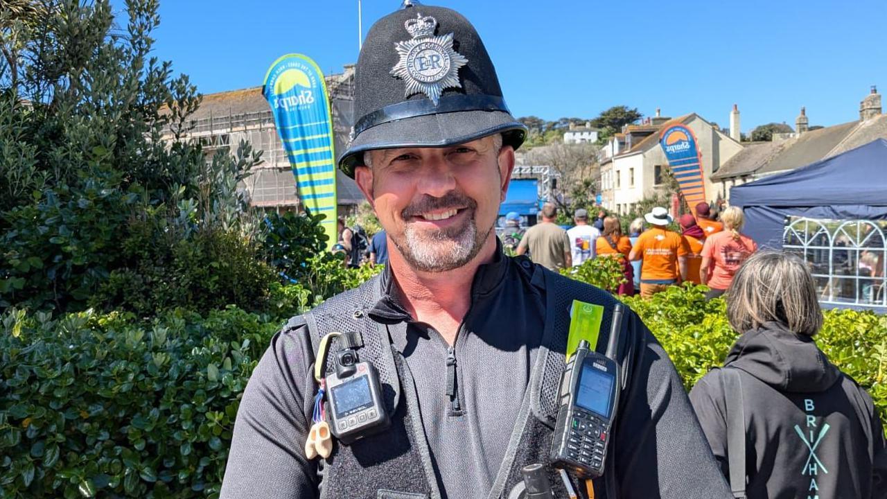 A UK police officer with a beard and traditional hat in black uniform with a number of devices attached.