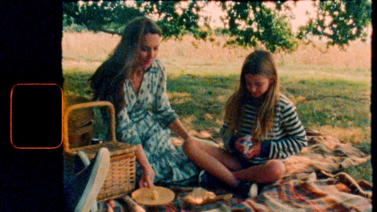 Catherine and Princess Charlotte sit together on a picnic blanket alongside food. Charlotte looks to be shuffling a deck of cards in the scene which is treated to appear like it is being played on old film