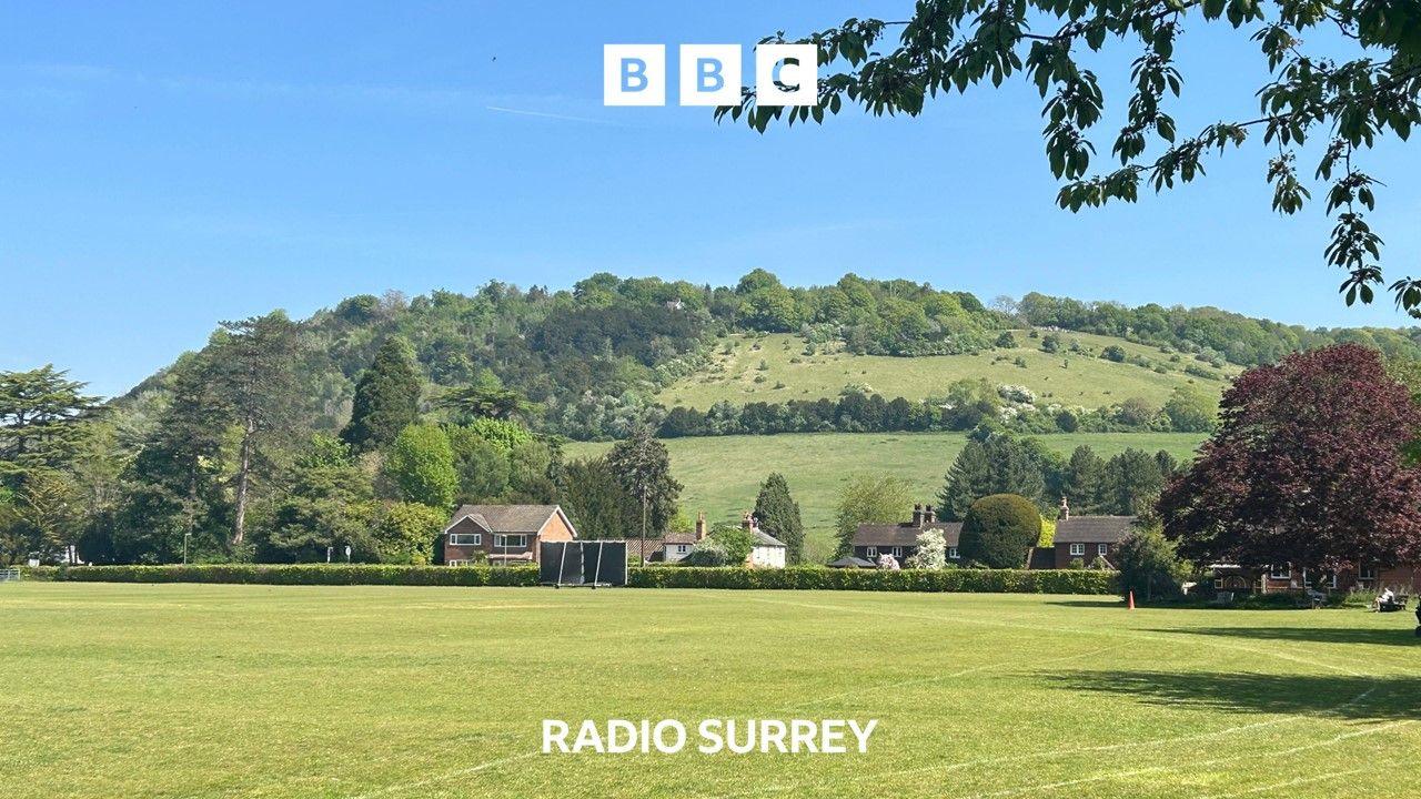 A hill rises in the background behind a neatly mown playing field and a number of houses. 