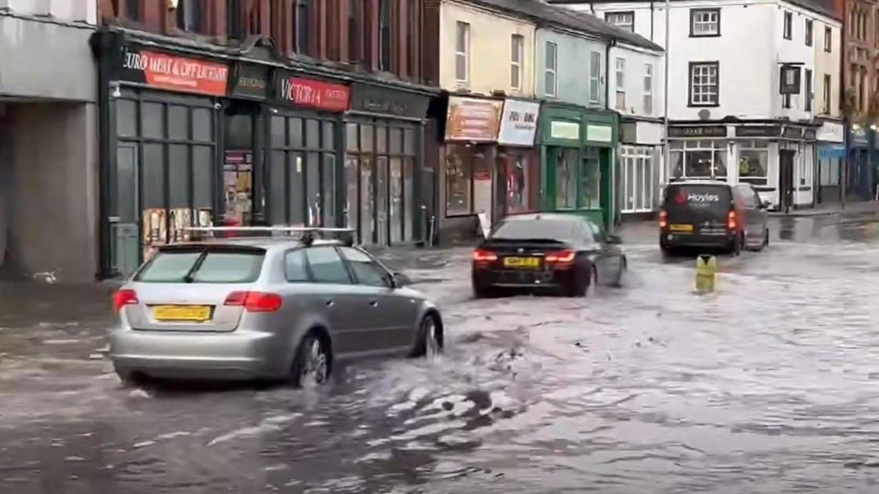 Cars drive through flooding on a road, with waters lapping up to the side of shops and buildings at the side of the road.