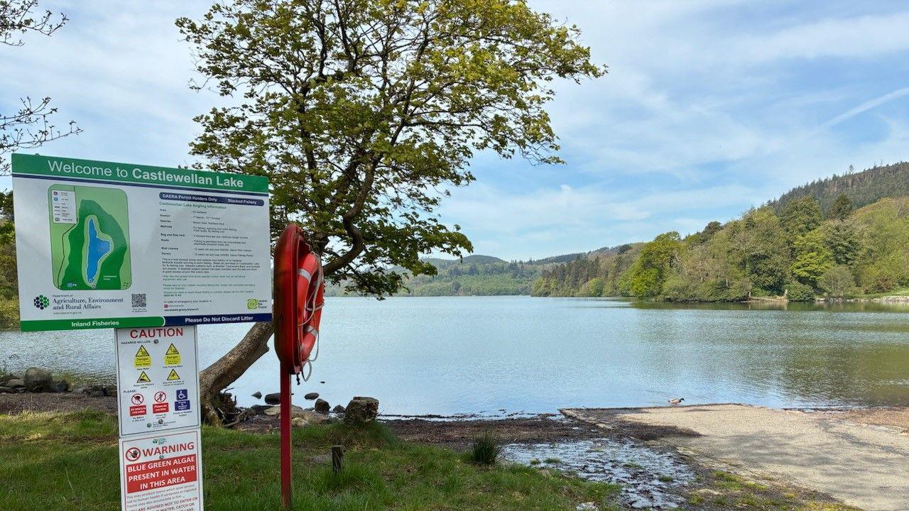 Castlewellan lake on a sunny day. The water is surrounded by trees with mountains in the background. A sign says 'Welcome to Castlewellan Lake'. Another sign shows a caution sign which is stood next to a life-ring.