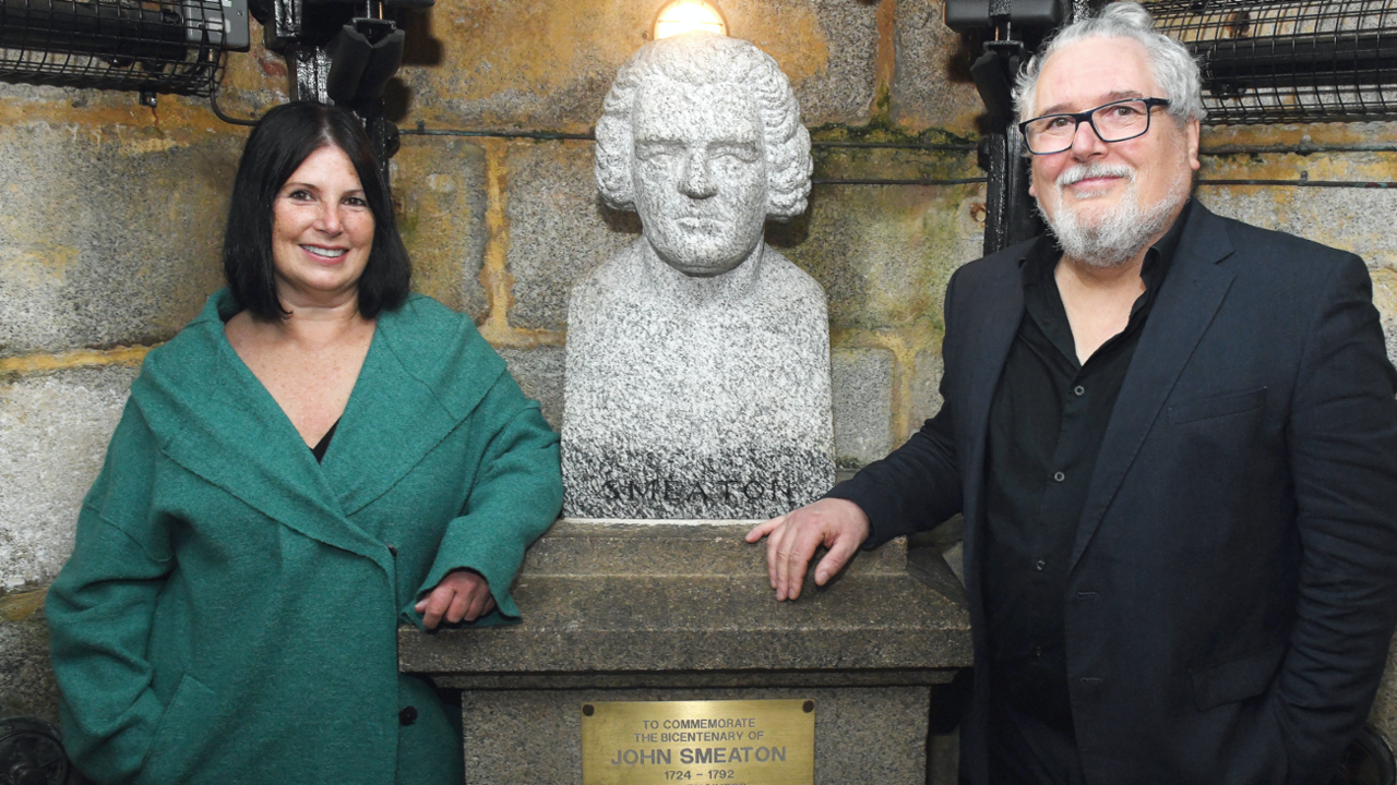 Miranda Housden (left) and Steve Conway (right) are pictured in front of the plaque. Miranda is wearing a long green jacket. She has short black hair. Steve is wearing a black shirt with a black blazer on top of it. He has grey hair and a grey beard with black glasses.