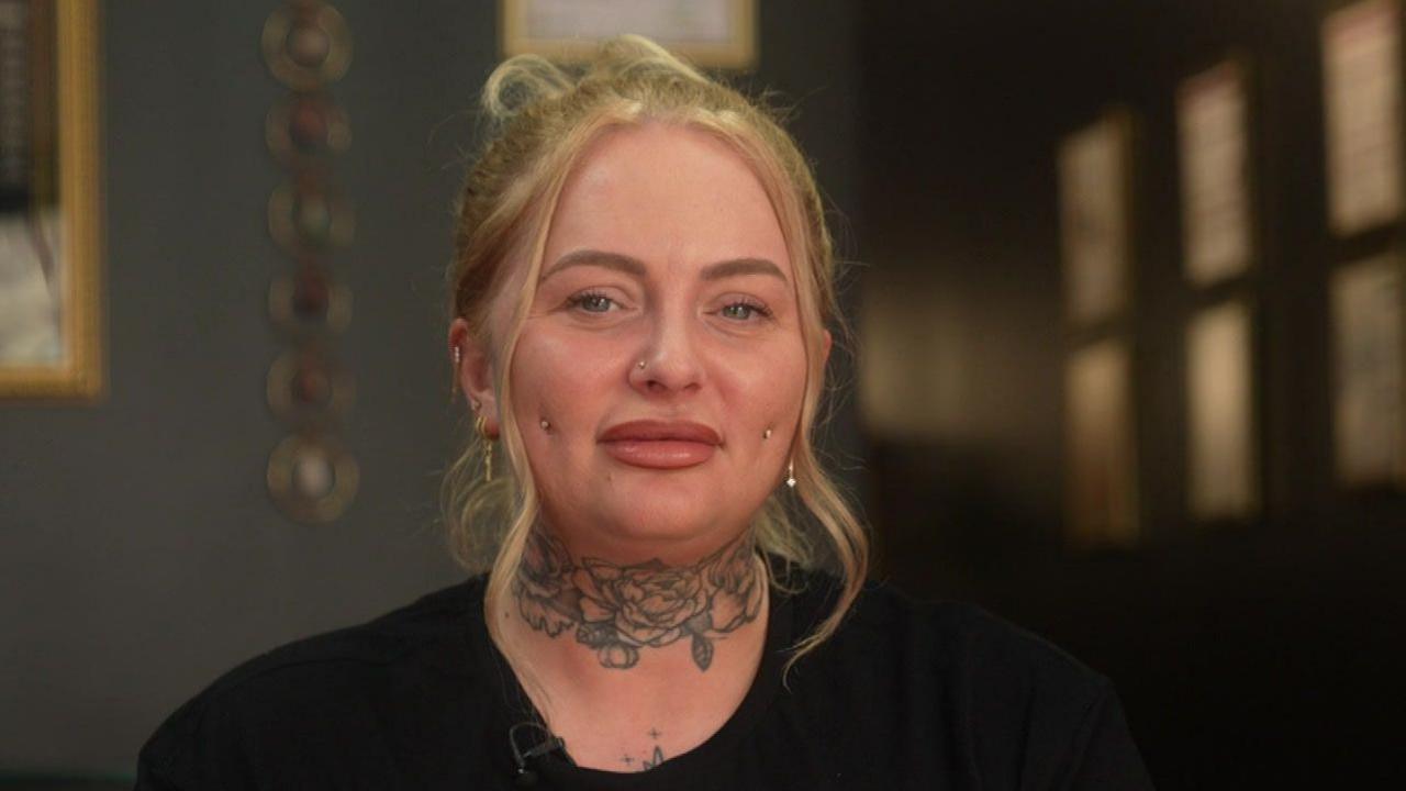 Lucy is sat in a professional piercing studio. Her blonde hair is tied up with two pieces loose, framing her face. She has a floral neck tattoo and piercings on each side of her cheeks and nose. She also has several ear piercings. She is wearing a black t-shirt with a logo on the front. An employee badge is place on the left side of her t-shirt.