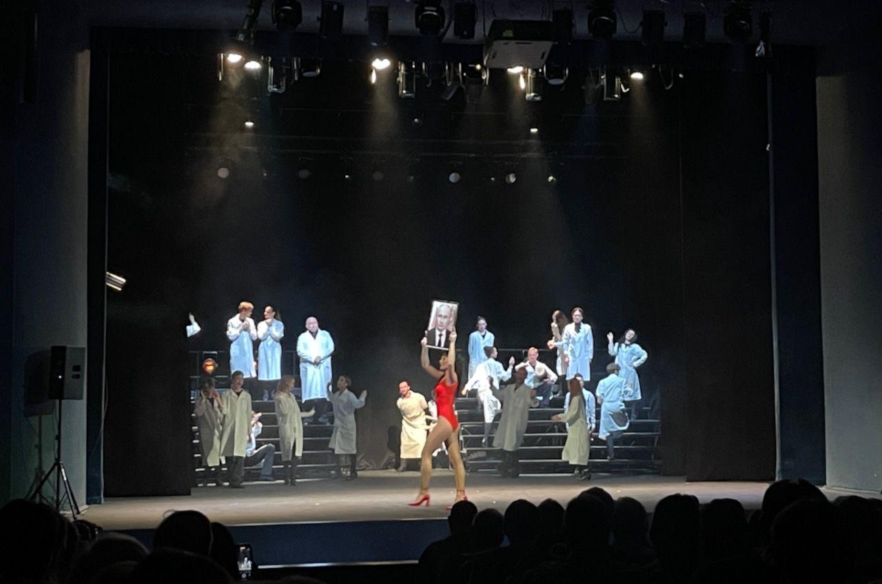 A woman in a leotard struts across a stage holding a large photo of Vladimir Putin above her head. Behind her are small crowds of performers sitting on tiered benches. 