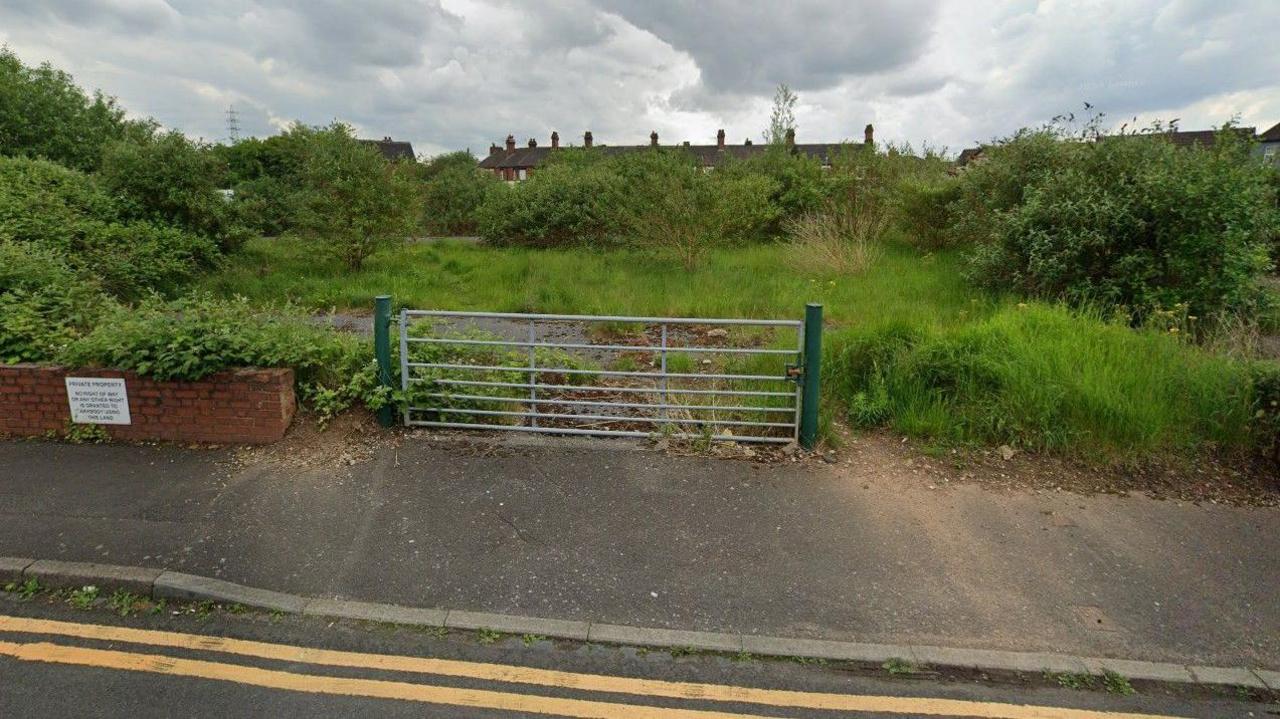 A patch of rough ground with grass and shrubs covering a hard-standing base. A metal gate and double yellow lines front the site, and the roofs of a row of terraced houses are visible behind.