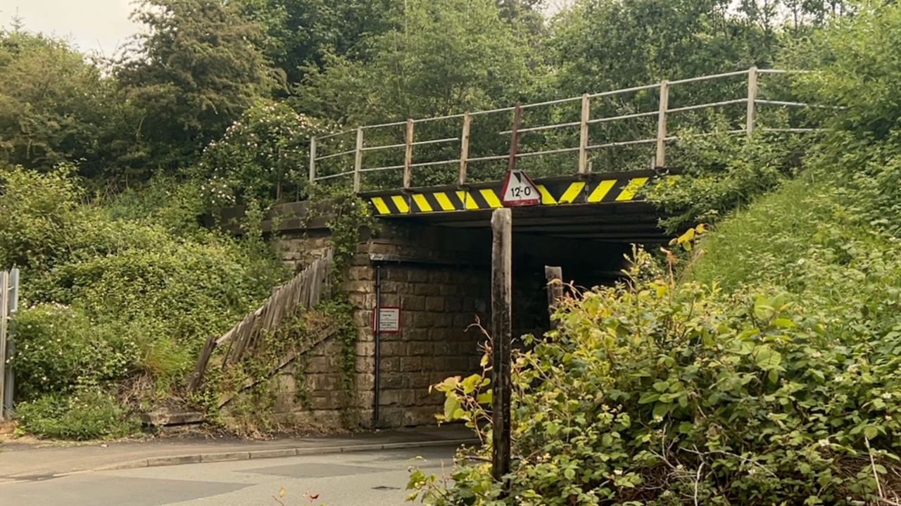 A former railway bridge that carried the Leamside Line is now overgrown with vegetation. There are a few pieces of metal missing, including from the railings, and there is a triangular sign warning people their vehicles must be under a certain height.