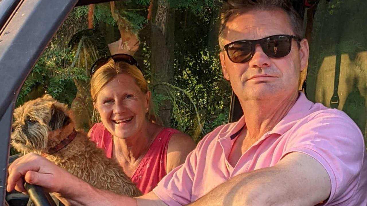 A man with sunglasses and pink shirt in a driving seat with a woman with blonde hair and red top in the passenger seat, and a small brown terrier between them
