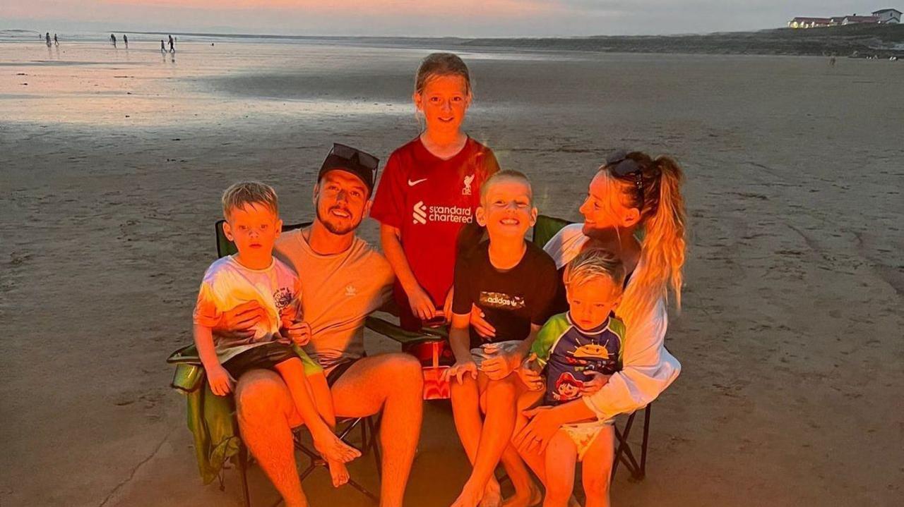 Nathan Osman and his partner Katie sitting on camping chairs with their four children at the beach. They are lit by the burning glow of the fire.