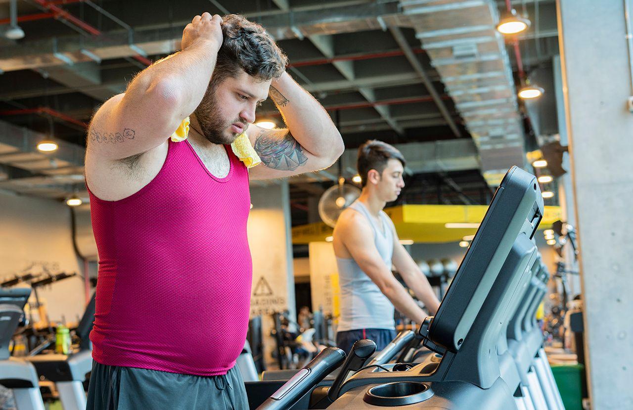 Two men on exercise machines in the gym, the man in foreground is using a flannel to wipe up sweat from working out, with his hands on his head.