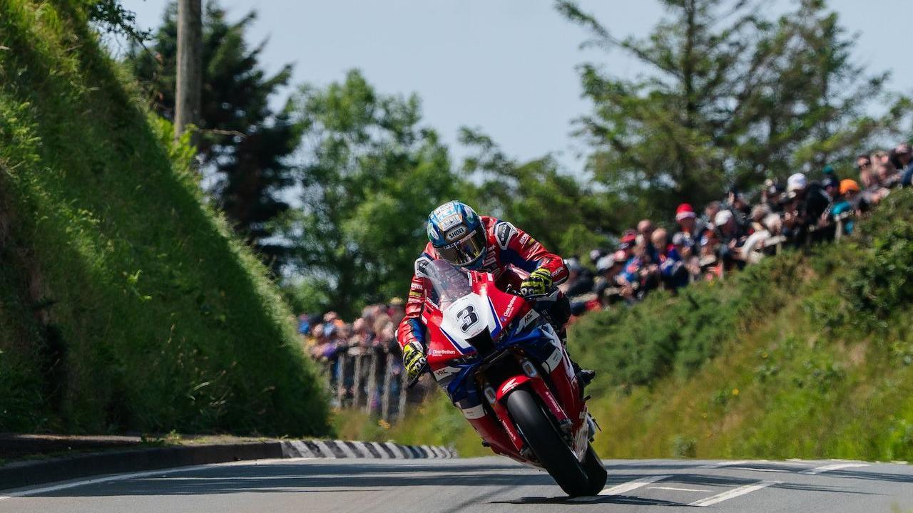 A rider on a red motorbike during the TT, there are fans on a grassy bank in the background.