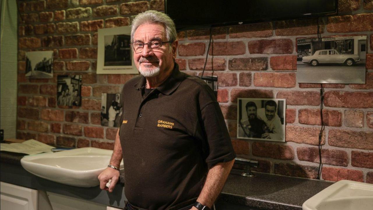 A man stands in front of two white sinks. The man is wearing a brown short-sleeved polo shirt and light grey hair and wears glasses