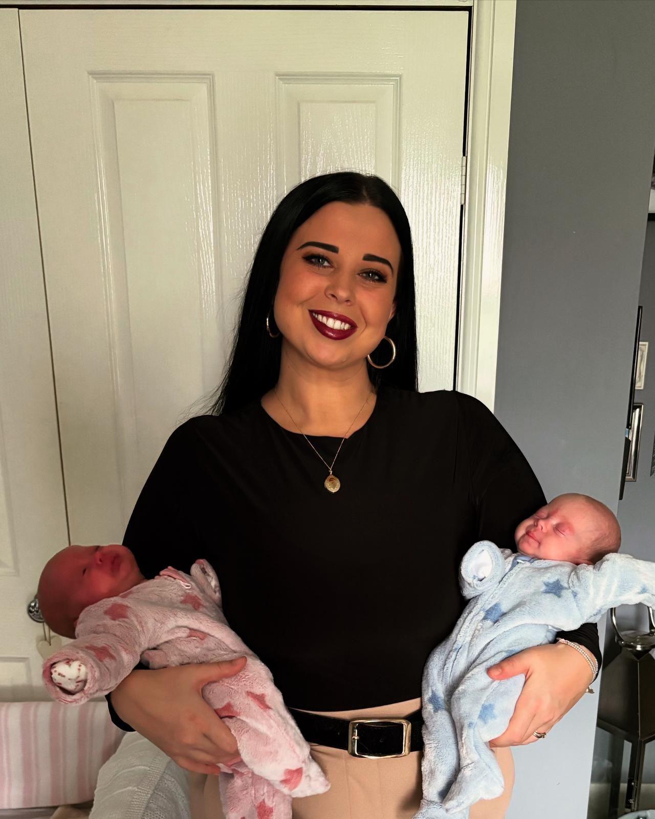 The picture shows a woman smiling at the camera. She's wearing a black long sleeved top and beige trousers. She has black hair and is wearing hooped earrings. She's holding a new born baby in each arm. One is wearing a pink baby grow, and the other is wearing a blue baby grow.