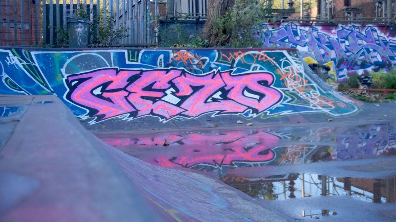 Colourful graffiti is painted on the ramps of a skate park. There are puddles on the ground and green bins in the background