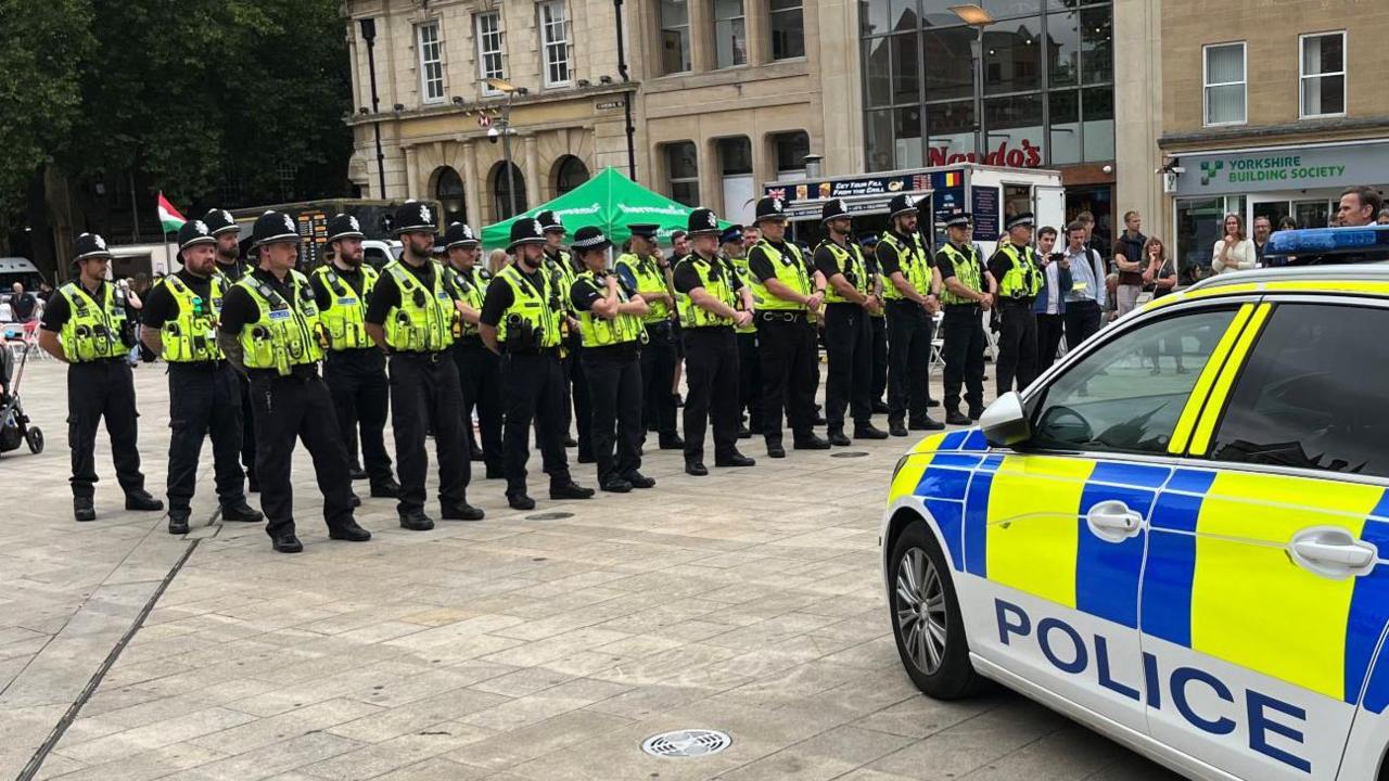 Line of police officers three deep stand in Peterborough City square. The are wearing hats and high-visibility waistcoats. A neon yellow-and-blue-chequered police car with the word POLICE on a door is parked close to the officers.