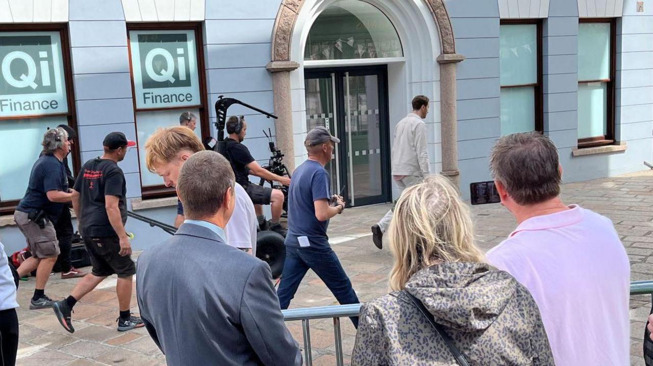 A film crew in the town of St Helier in Jersey watched by members of the public and police. An actor walks down a shopping street, followed by various members of the crew including a mounted camera operator.