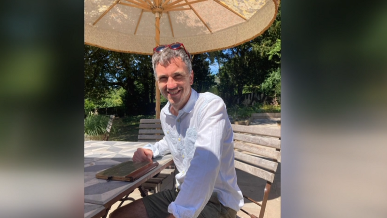 James Palmer-Bullock wearing a white shirt and grey shorts, sitting outside under some sunny weather.