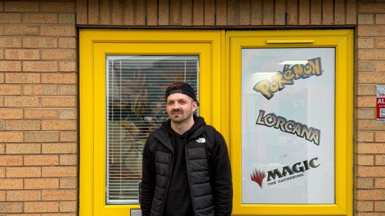 Man in a backwards baseball cap, with a stubble beard and moustache. He is wearing a black hoodie and a black gilet and is standing outside a shop with yellow PVC doors and windows, 