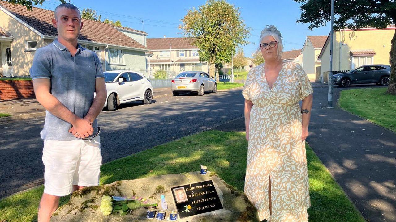 Jason Arksinon, on the left and his mum Kathleen standing beside a memorial stone to Arlene Arkinson. Jason is wearing a blue shirt and shorts, Kathleen, with her arms at her side, is wearing a patterned dress