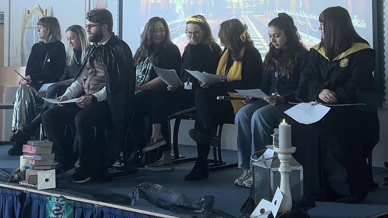 Eight teachers sitting on a stage at the front of a school hall. They are all wearing Harry Potter-themed fancy dress.