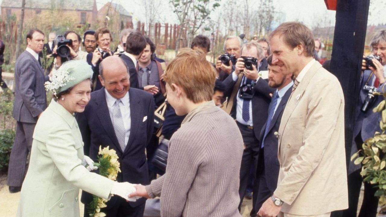 Theo Gay Anderson, who has red hair and is wearing a light brown cardigan, shakes the white-gloved hand of Queen Elizabeth II who is wearing a light green outfit with a matching hat.