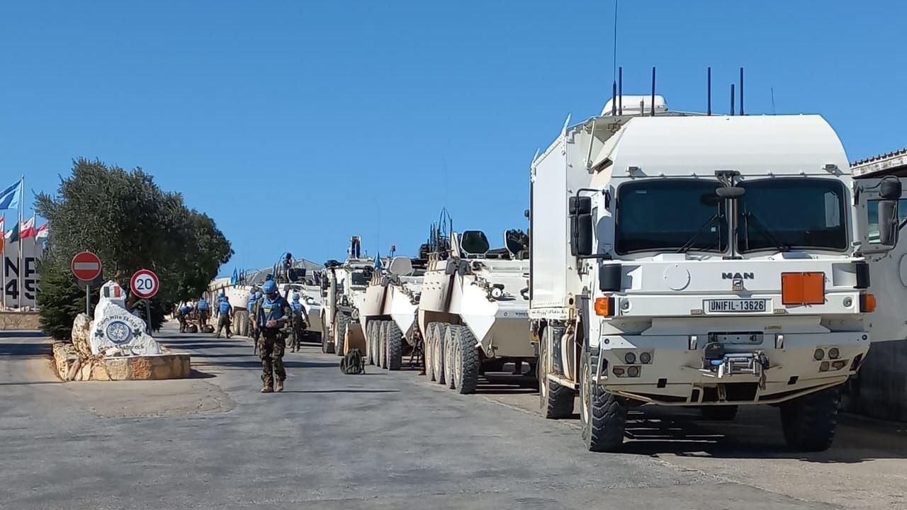 A convoy of UN vehicles recently stationed at an entrance to Camp Shamrock.  The large white vehicles are parked in a line and a group of soldiers wearing camouflage uniforms, blue UN helmets and flak jackets are preparing to depart for an operation.   There is a sign at the entrance to the camp which says "Céad Míle Fáilte" - the Irish words for "One hundred thousand welcomes".  The sign is decorated with the Irish flag and two shamrocks. 