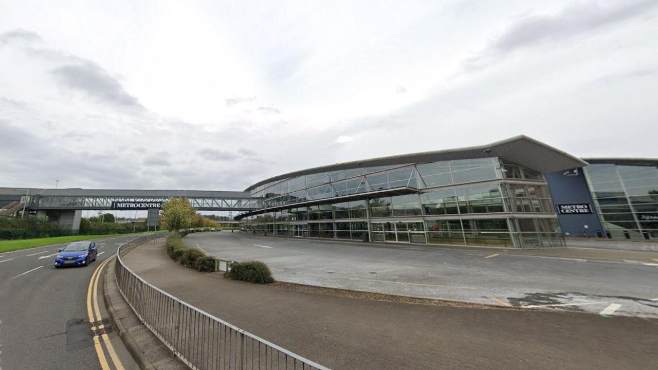 A picture of the Metrocentre bus station. The building is large and made of glass. A bridge passes over the a road leading to a train station. A sign displaying the shopping centre's name is displayed on the bridge.