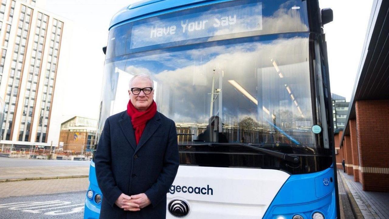 A man with a red scarf stands in front of a large bus. He has a blue coat on, black-rimmed glasses and a slight smile beneath short white hair. The bus has the last letters of "Stagecoach" visible and is blue and white with large glass windows and a digital sign saying: "Have your say".