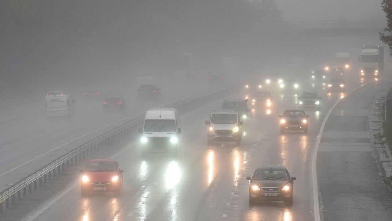 a grey, dull and wet motorway showing a dozen cars driving with their lights on in the gloomy conditions, with a motorway bridge in distance